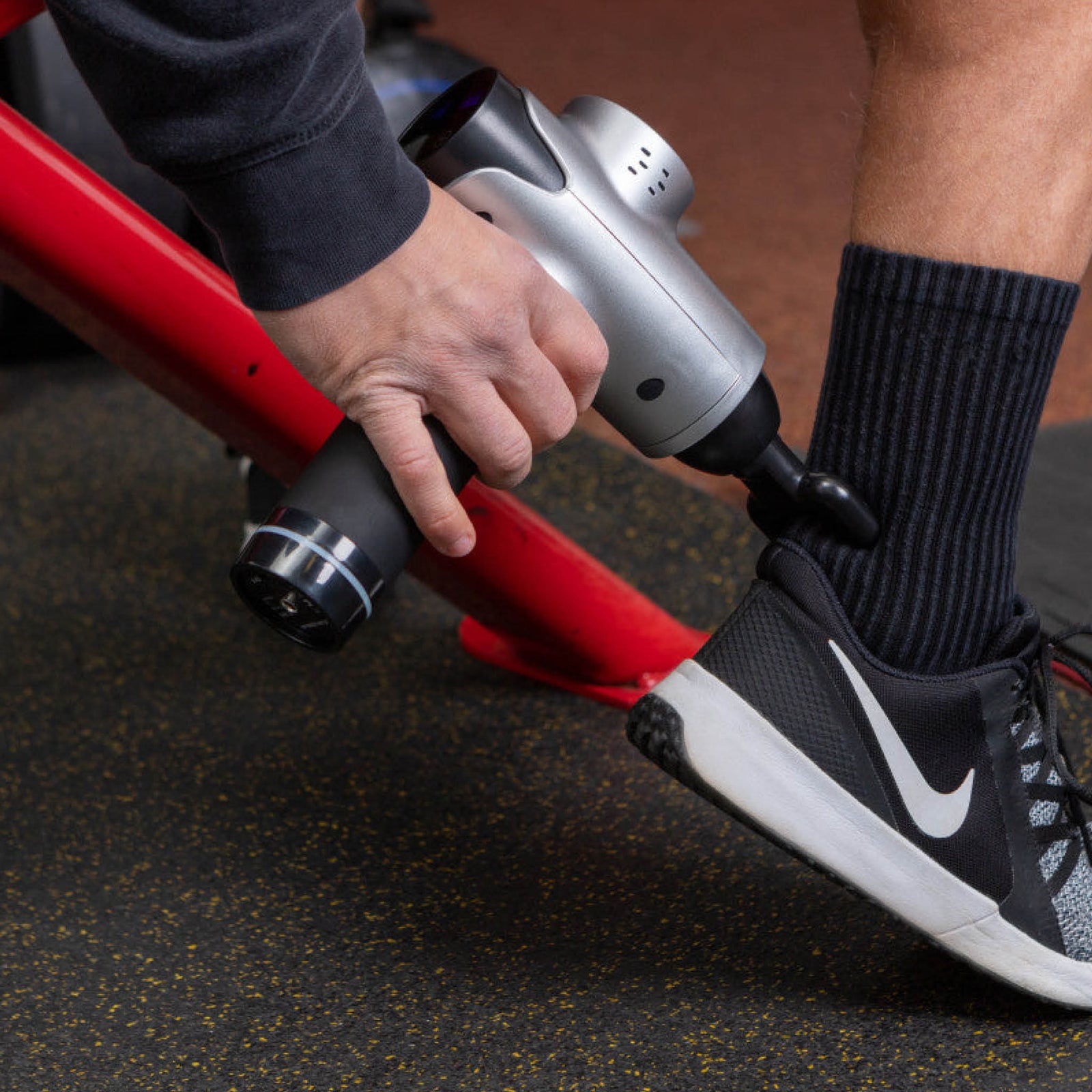 Seated on red gym equipment, a person uses the Reviber Massage Gun by Reviber for deep-tissue massage on their ankle while wearing black Nike sneakers and black socks.