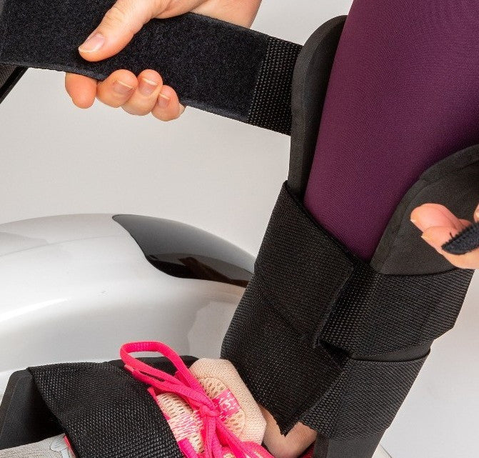 A person in a dark purple outfit fastens black Reviber Mini Bike Pro Leg Supports (one pair) by Reviber around their leg and foot, which is in a bright pink sneaker—likely for use with a pedal exerciser.