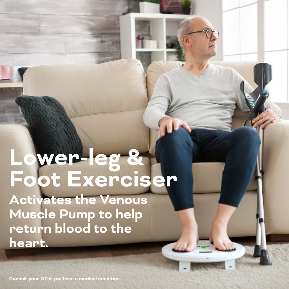 An older man sits on a beige sofa using crutches, resting his bare feet on the Reviber Circulation Stim by Reviber, an electronic muscle stimulator that boosts lower leg strength and helps return blood to the heart by activating the venous muscle pump.