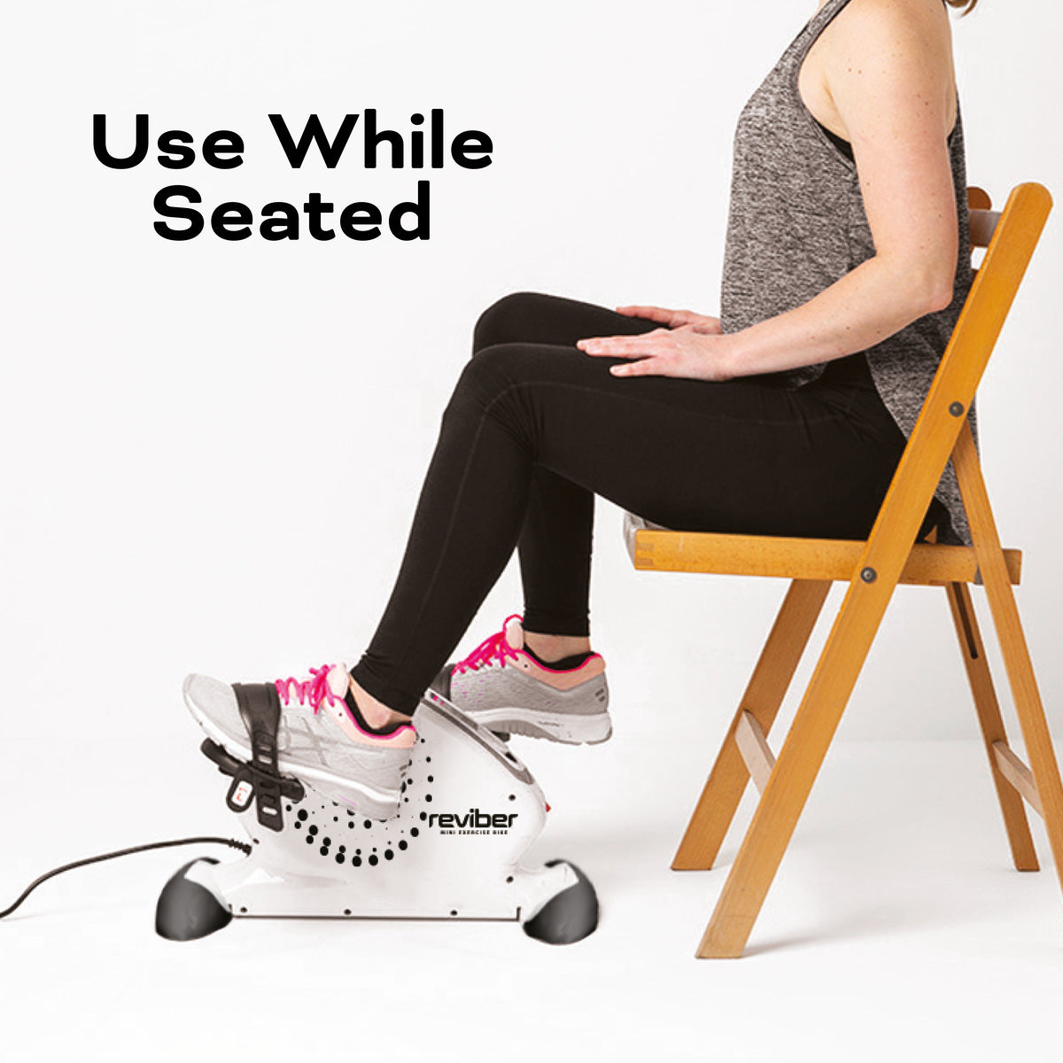 Wearing athletic clothes and sneakers, a person sits on a wooden chair using the Reviber Mini Motorised Exercise Bike by Reviber. Text: Use While Seated—ideal for low-impact rehabilitation.