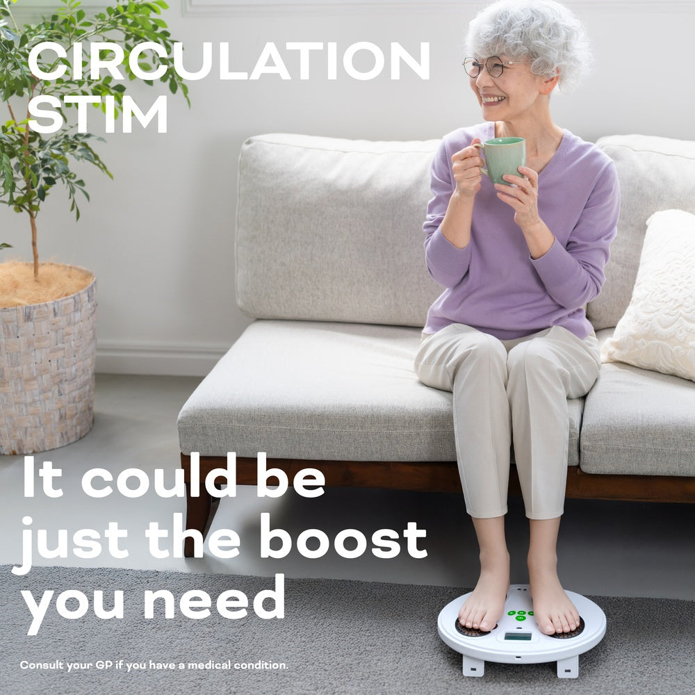 An older woman with short gray hair sits on a sofa, smiling and holding a mug, while using the Reviber Circulation Stim - Electronic Muscle Stimulator by Reviber. The text reads: CIRCULATION STIM. It could be just the boost you need.