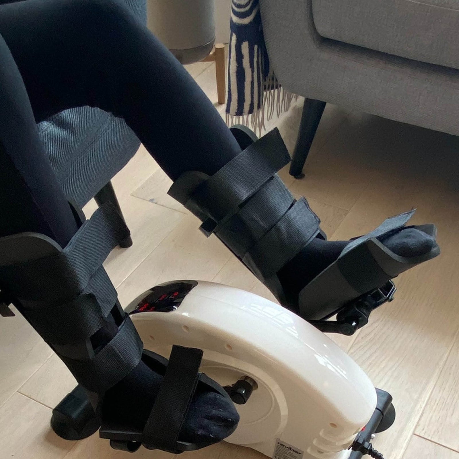 A person uses the Reviber Mini Bike PRO by Reviber, a dual-function motorised and manual resistance exerciser, while seated indoors on a chair—ideal for low-impact rehabilitation, even with black medical braces on both legs.