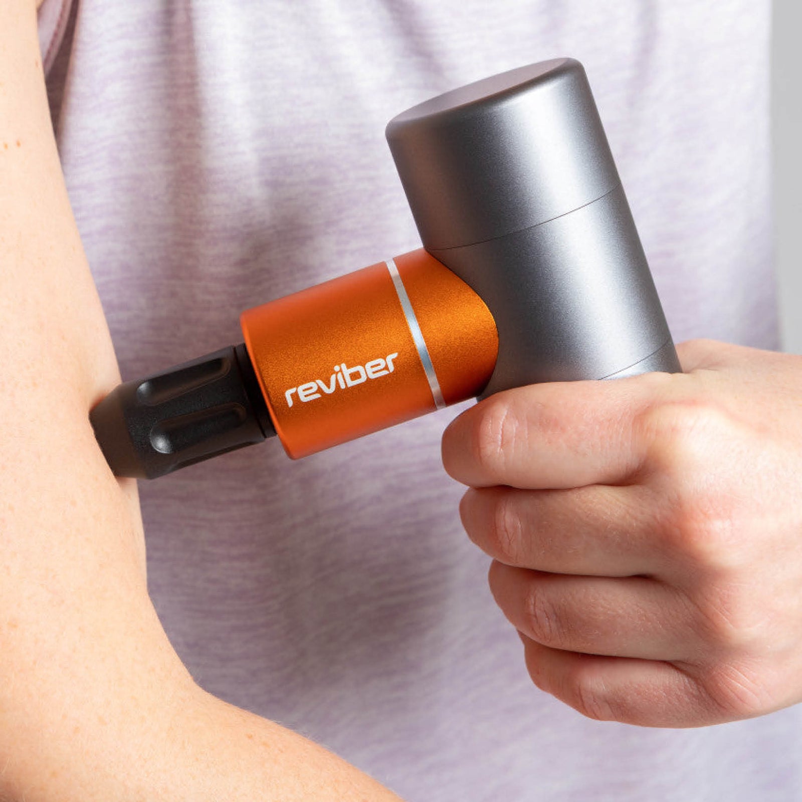 A person wearing a light purple shirt uses the Reviber Mini Massage Gun With Heat Therapy, holding it firmly on their upper arm for a deep tissue massage. The orange and gray handheld device is from Reviber.