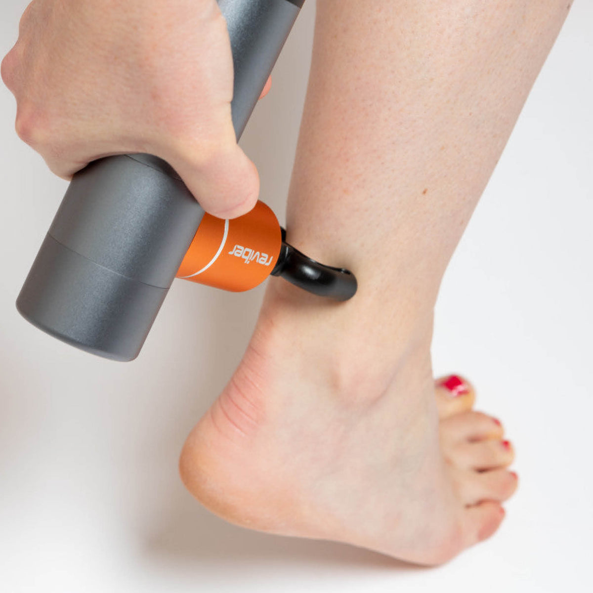 A person uses the Reviber Mini Massage Gun With Heat Therapy by Reviber on their lower leg near the ankle. Their toenails are painted red, and a plain white background highlights this relaxing massage moment.
