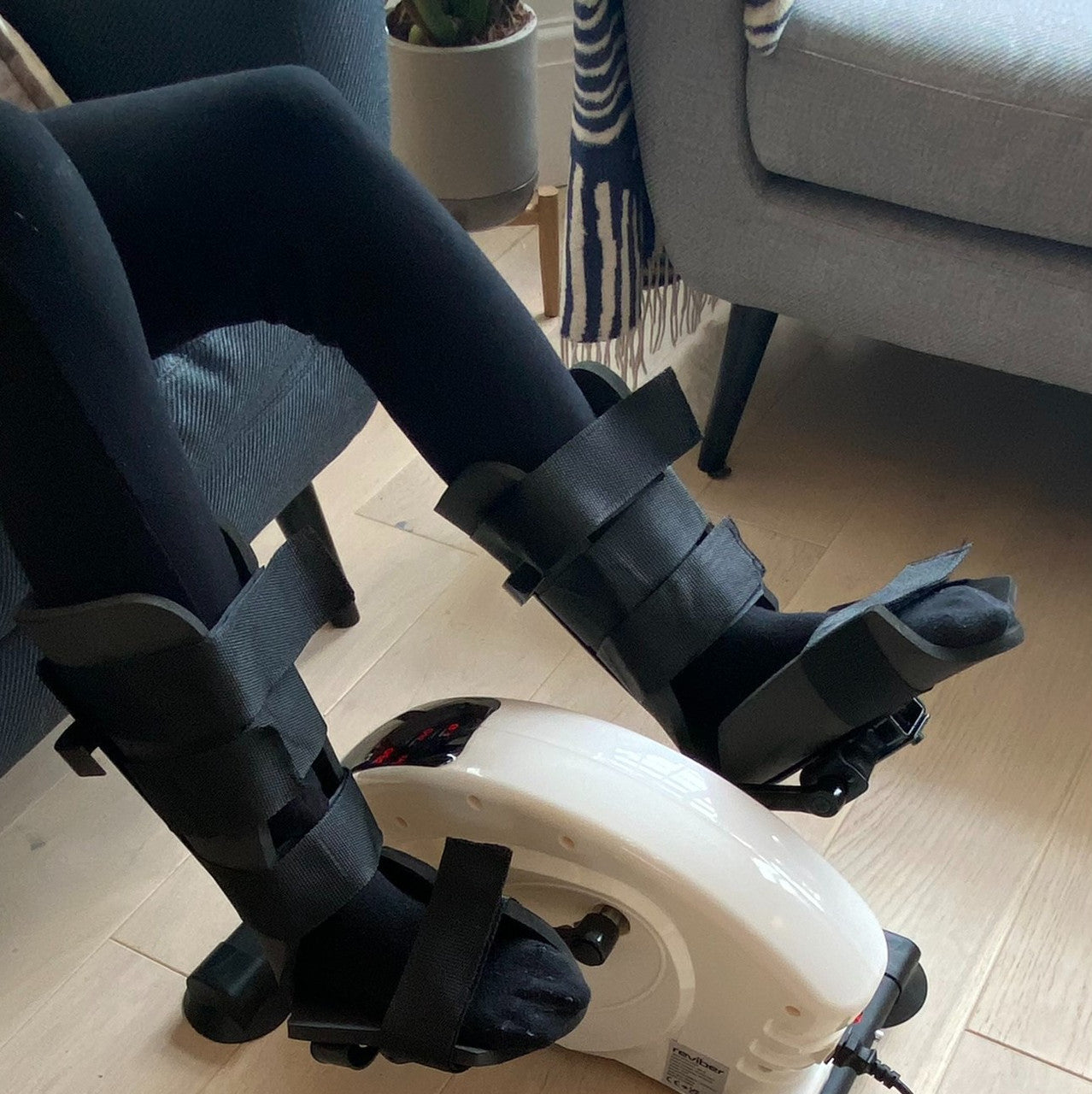 A person with Reviber Mini Bike Pro Leg Supports (one pair) by Reviber and black medical braces pedals a compact exerciser indoors, placed in front of a gray chair and a plant on a wooden floor.